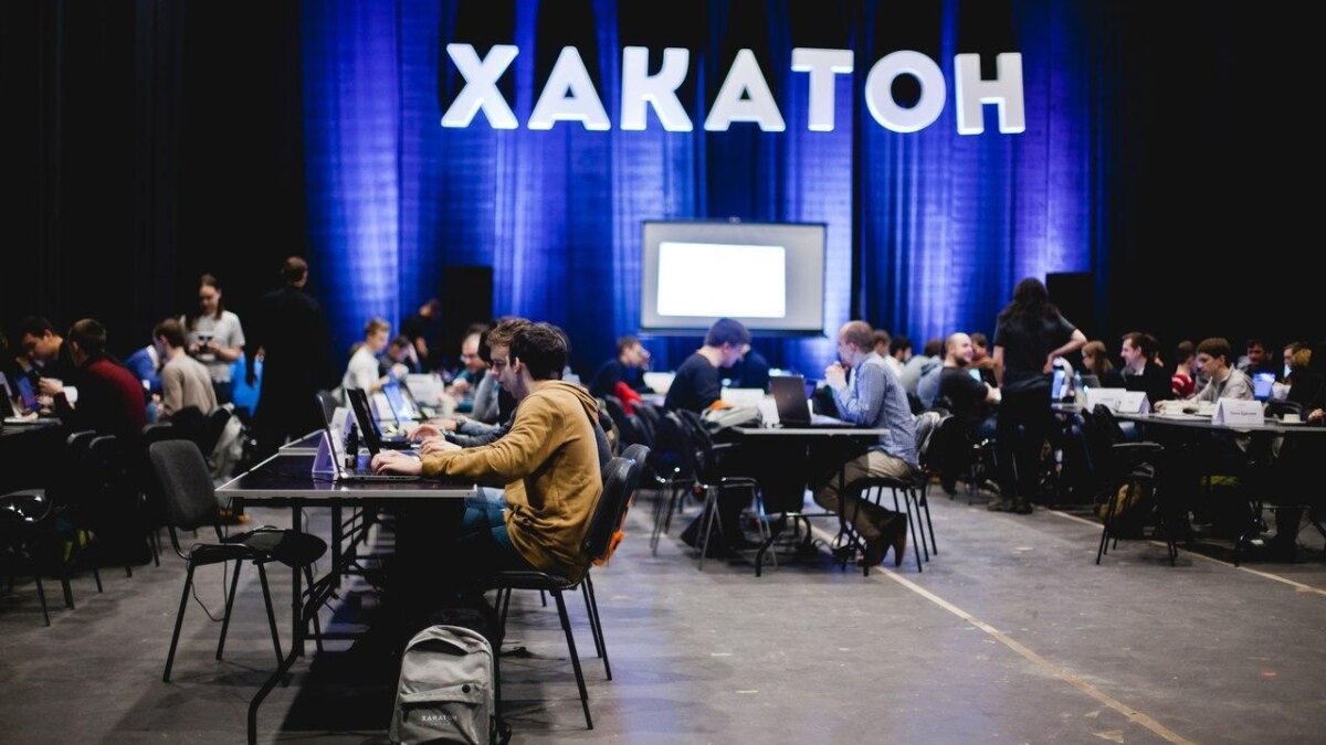Участники хакатона за ноутбуками на сцене с экраном и большим знаком Хакатон