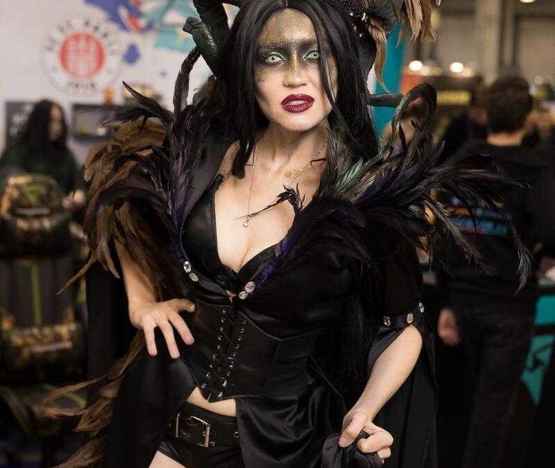 Женщина в черном костюме с крупной шляпой из перьев и украшением на голове на стенде ИгроМир 2018