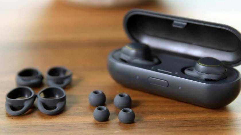 На изображении показаны беспроводные наушники Samsung Galaxy Buds в кейсе и дополнительные насадки на деревянной поверхности