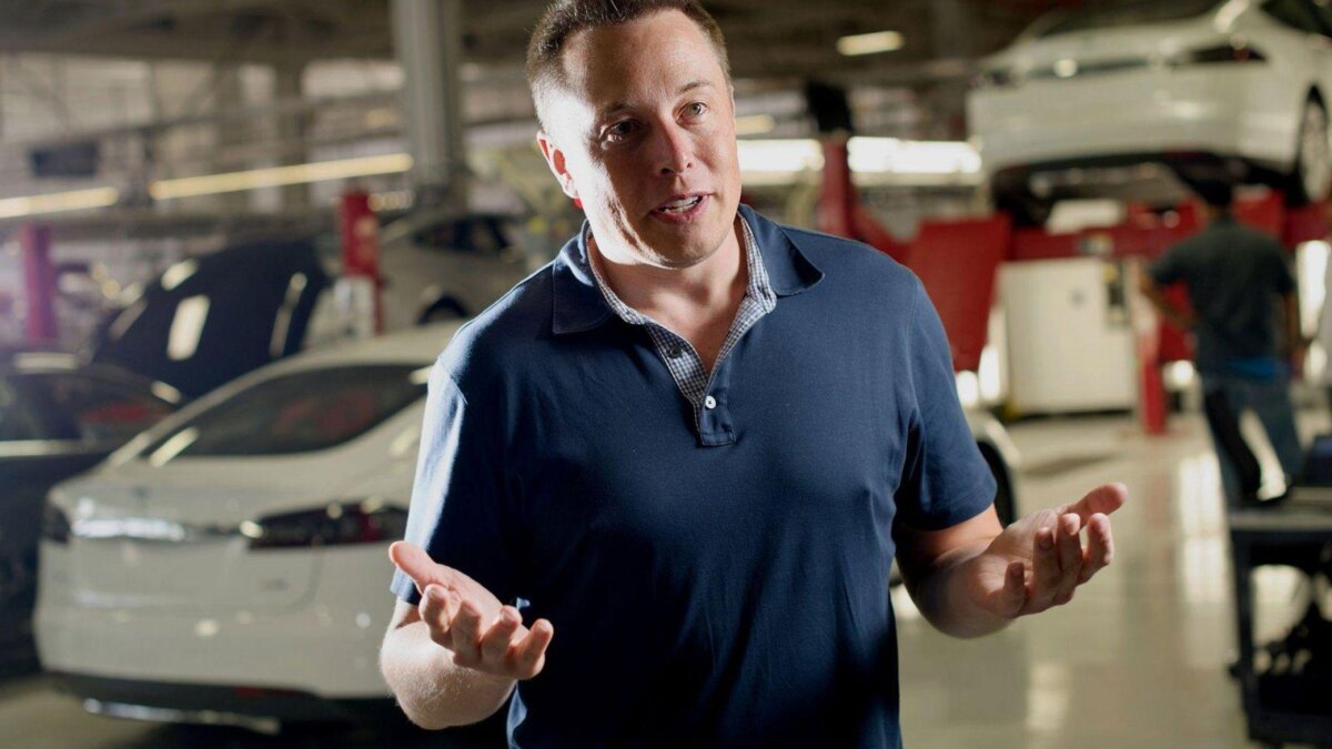 Человек объясняет что-то в автомастерской с автомобилями и подъемниками на заднем плане