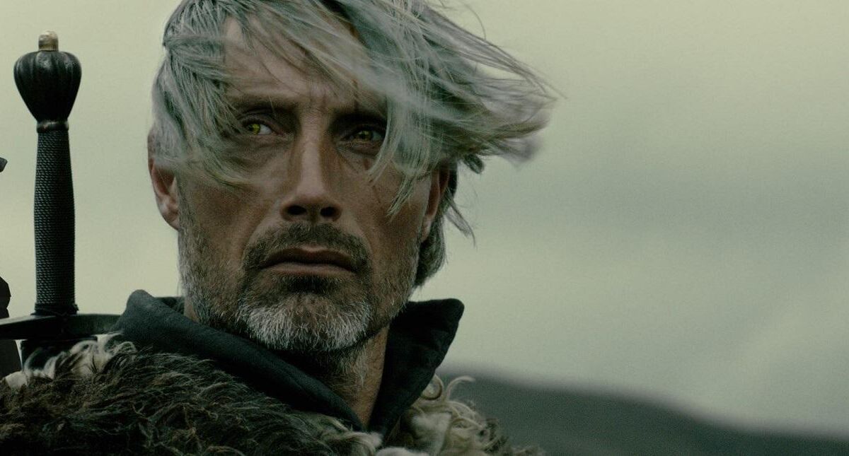 Портрет Мадса Миккельсена с длинными светлыми волосами и бородой в темной одежде на фоне неба