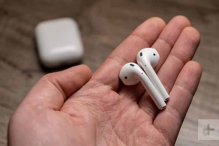 A pair of Apple AirPods in a hand with their case in the background. 