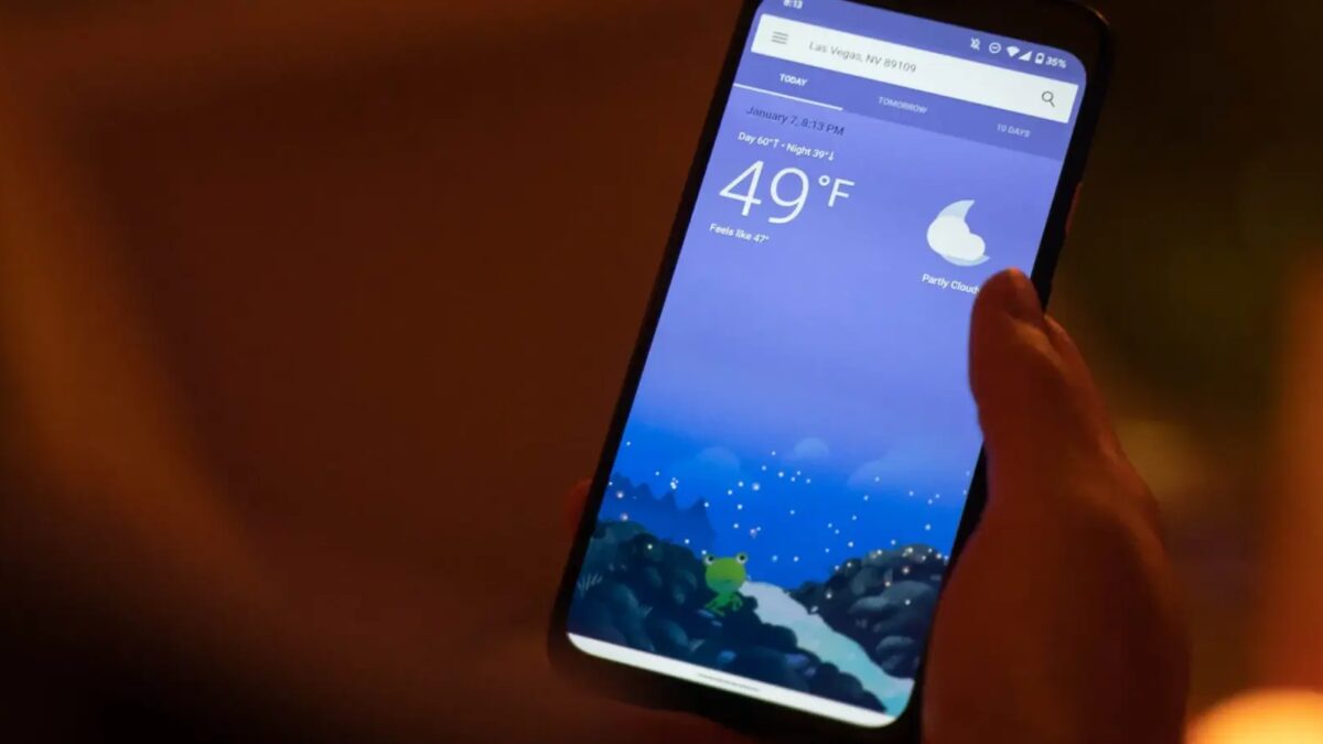 Экран смартфона с приложением погоды на фоне темных с голубым облаком и фифером