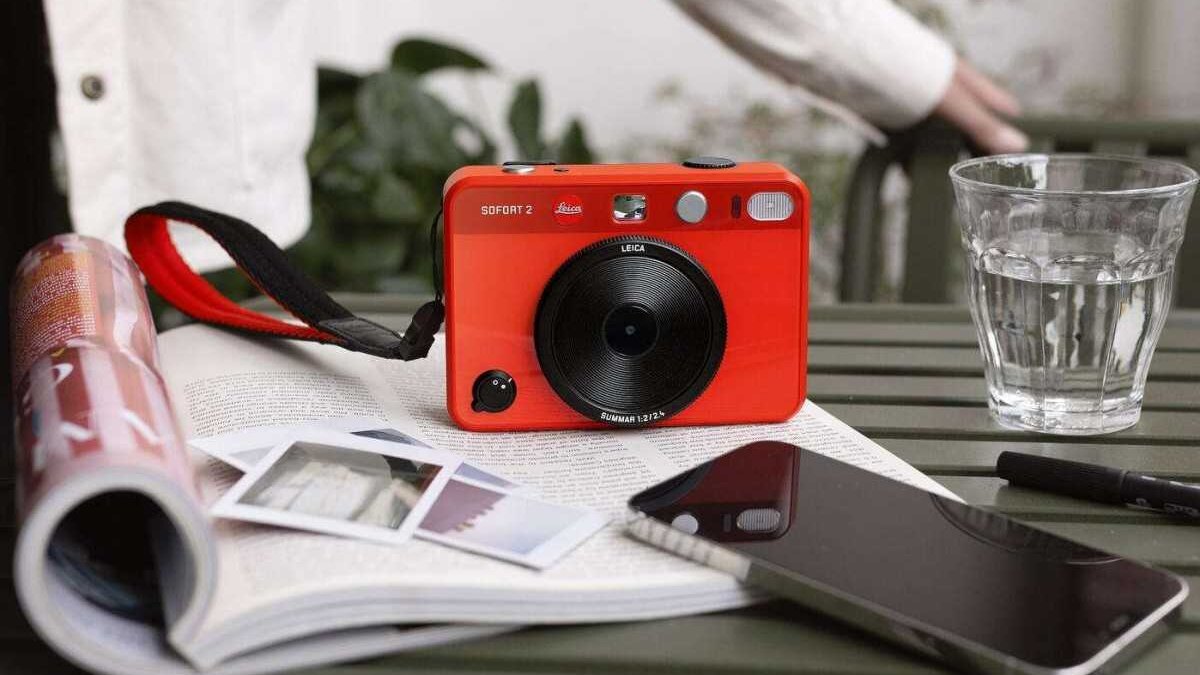 Красная камера Leica Sofort 2 лежит на столе рядом с телефоном и водой