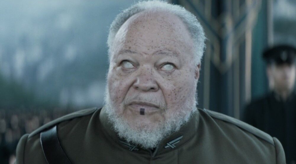 Портрет пожилого мужчины с бледной кожей и белой бородой в военной форме в окружении людей и охраны