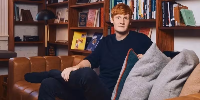Молодой человек сидит на диване в домашней библиотеке среди книг и декоративных подушек