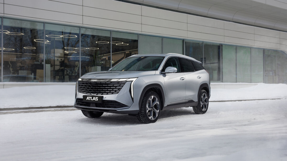 Новая переднеприводная версия кроссовера Geely Atlas серебристого цвета на зимней дороге на фоне современного здания