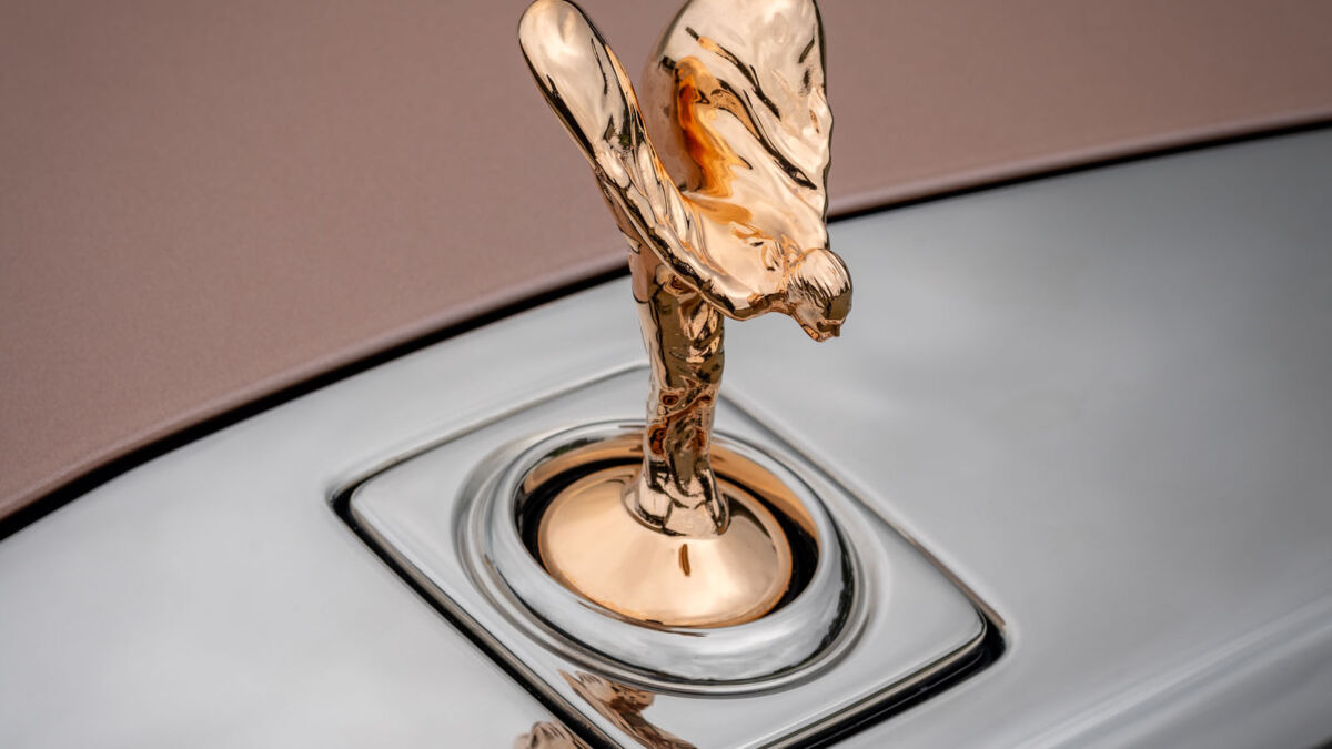 Золотая сувенирная статуэтка Rolls-Royce «Дух экстаза» на фоне логотипа бренда